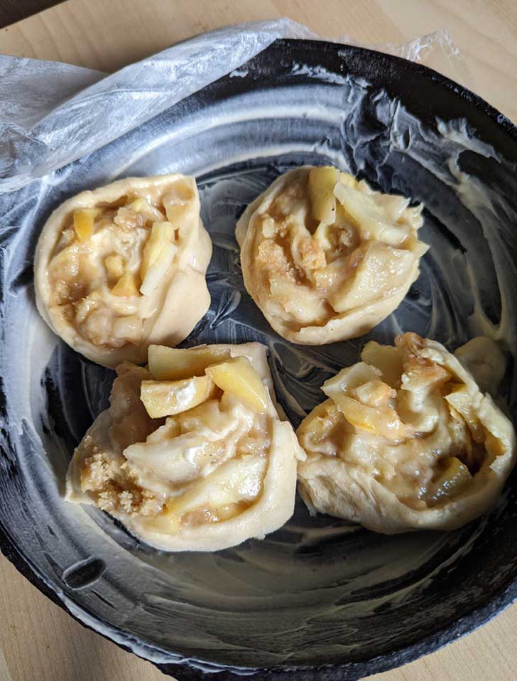 Unbaked cinnamon rolls with a filling of apple pie in a buttered cast iron skillet.