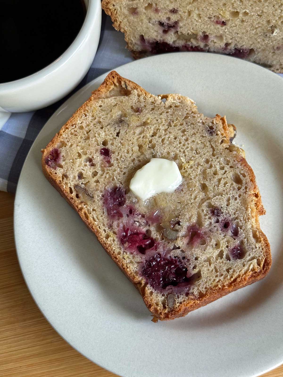 A slice of lightly toasted blackberry quick bread on a plate with butter and coffee.