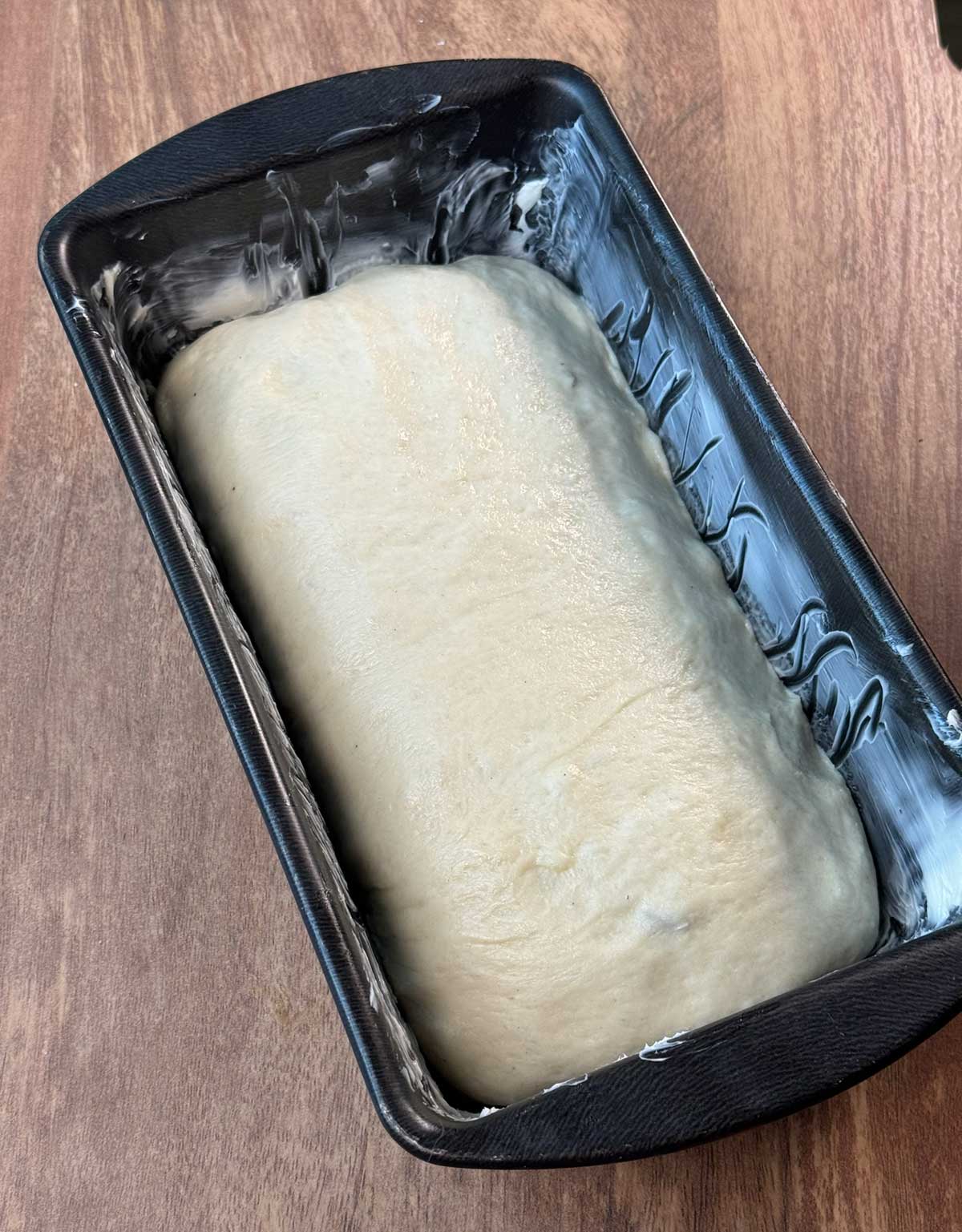 White bread dough in a black loaf pan greased with butter.