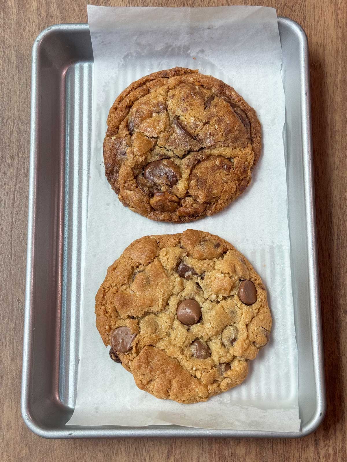 Two large chocolate chip cookies on a mini sheet pan. The first one has grated milk chocolate in it.
