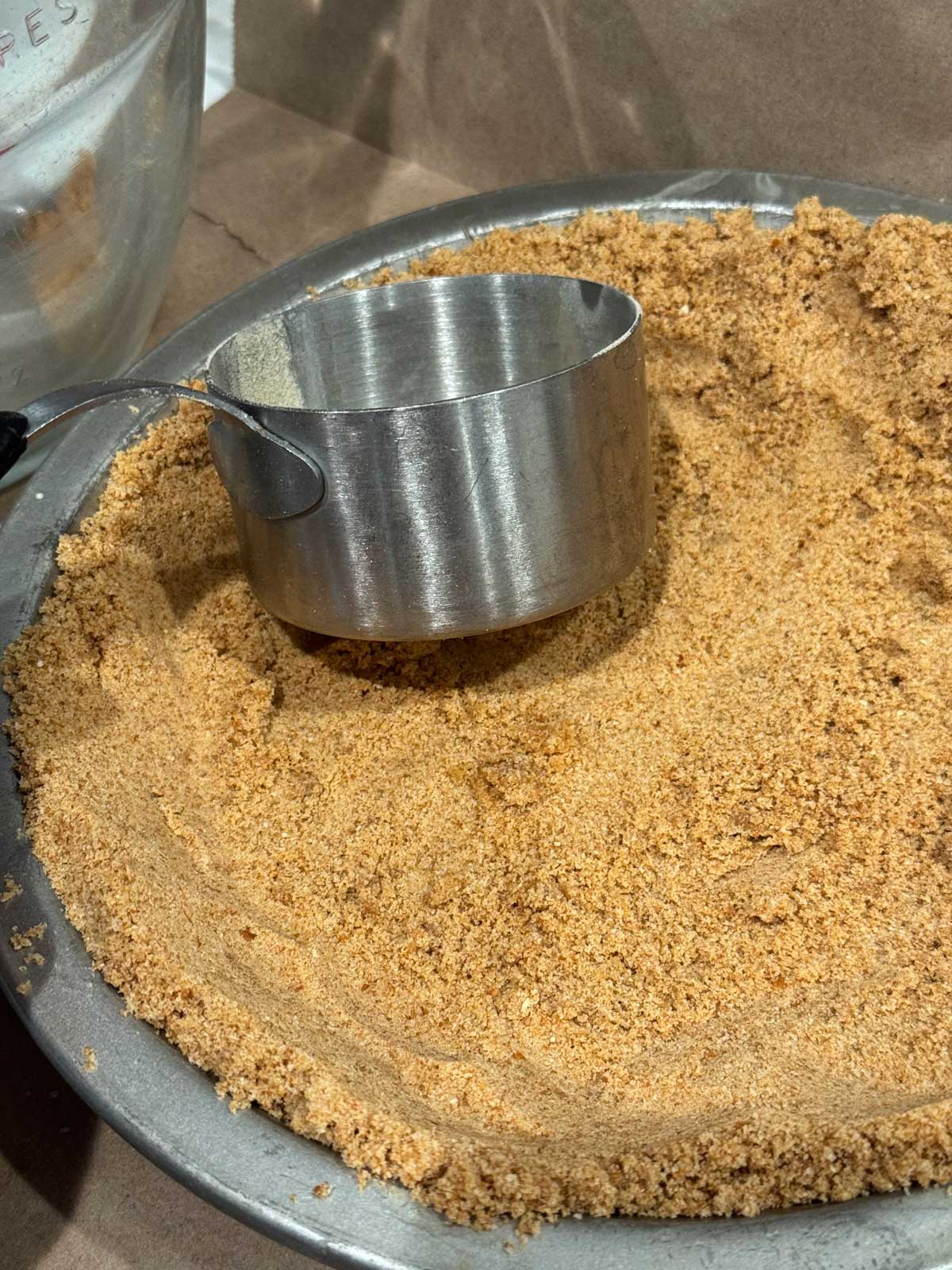 Pretzel crust being pressed into a metal pie pan with the side of a one cup measure.